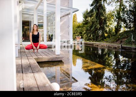 Donna mid adulta meditating mentre accovacciandosi sul pavimento di legno vicino stagno Foto Stock