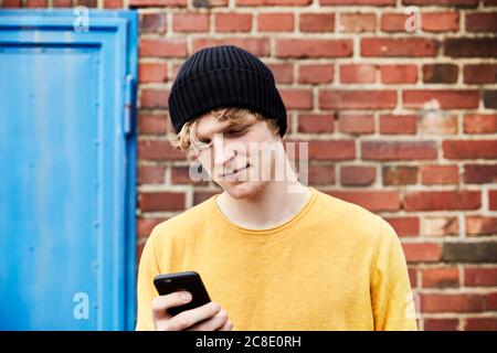 Ritratto di un giovane uomo che indossa il cappuccio e guarda lo smartphone all'interno fronte di muro di mattoni Foto Stock