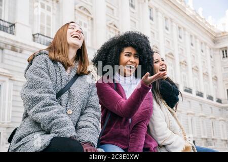 Amici multietnici che ridono mentre si siedono contro il Palazzo reale di Madrid, Spagna Foto Stock