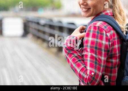 Primo piano di una donna sorridente che indossa una maglietta controllata con zaino mentre in piedi sul ponte Foto Stock