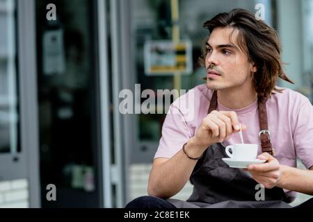 Premuroso proprietario di sesso maschile che tiene una tazza di caffè mentre si siede all'esterno della caffetteria Foto Stock