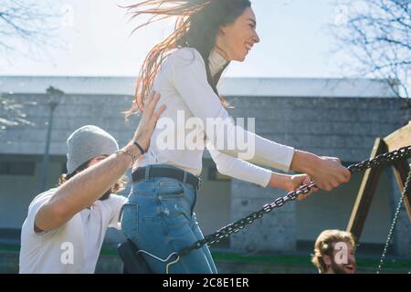 Giovane uomo che spinge amici giocosi che oscillano nel parco durante il sole giorno Foto Stock