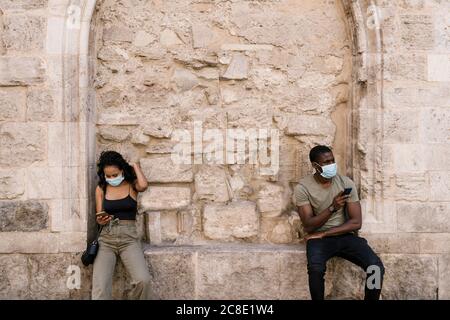 Uomo e donna indossano maschere mentre si utilizzano smartphone contro parete Foto Stock