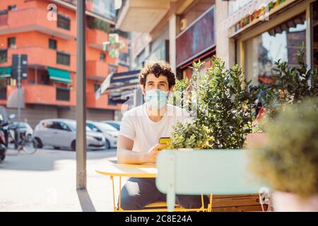 Un giovane uomo premuroso che indossa una maschera seduto al bar sul marciapiede città Foto Stock