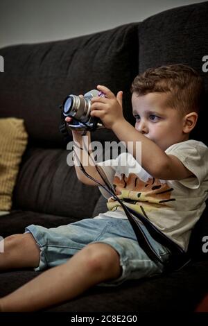 Ragazzo carino fotografando con la macchina fotografica mentre si siede sul divano a. casa Foto Stock