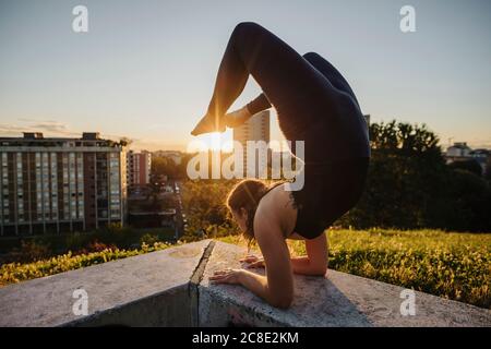 Giovane donna flessibile che esegue yoga sulla parete di ritegno in città durante il tramonto Foto Stock