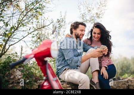 Accoppia usando lo smartphone mentre sei seduto su una parete di ritegno cielo Foto Stock