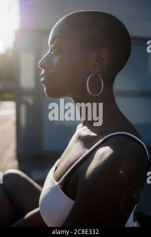 Primo piano di una giovane donna premurosa con la testina rasata che guarda lontano mentre si è seduti all'aperto durante la giornata di sole Foto Stock