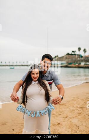 Coppia in attesa felice che tiene il cartello del nome mentre si sta in piedi sulla spiaggia, Cascais, Portogallo Foto Stock