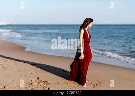 Donna delicata in abito rosso che cammina al mare, sentendo il sole Foto Stock