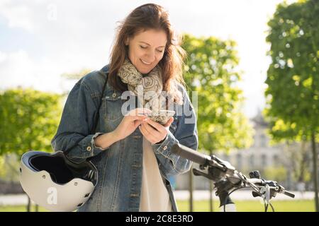 Donna sorridente con casco e bicicletta utilizzando smartphone mentre situato nel parco cittadino Foto Stock