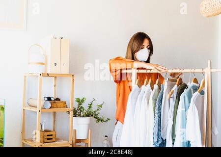 Donna stilista di moda che lavora a casa con stativo di vestiti che indossa maschera protettiva Foto Stock