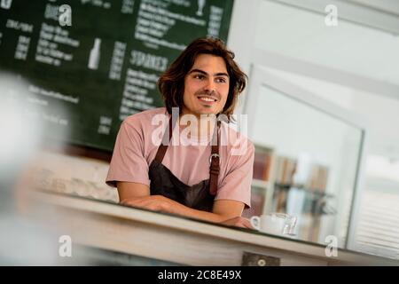 Sorridente uomo proprietario guardando via mentre si sta in piedi al bancone dentro caffetteria Foto Stock