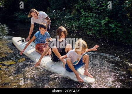 Padre che spinge i ragazzi che spruzzano acqua mentre si siede sul paddleboard dentro streaming Foto Stock