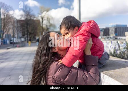 Affettuosa donna che bacia sua figlia seduta su un muro di ritegno città Foto Stock