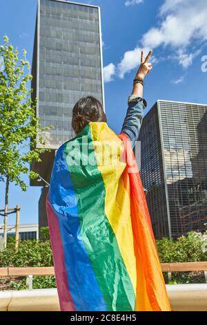 Donna gesturing segno di pace mentre porta la bandiera arcobaleno in città Foto Stock