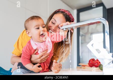 Primo piano di madre sorridente che tiene simpatica bambina giocando con rubinetto in cucina Foto Stock
