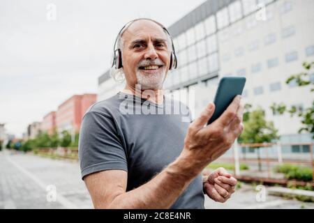 Uomo anziano sorridente che indossa le cuffie utilizzando lo smartphone mentre si è in piedi in città Foto Stock