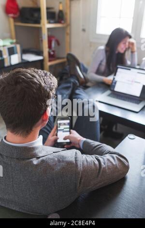 Uomo d'affari rilassato con smartphone alla scrivania e donna in background in funzione Foto Stock