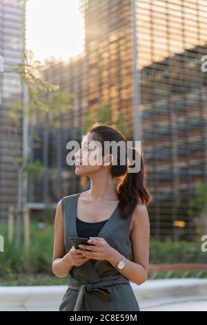 Donna d'affari premurosa che tiene lo smartphone mentre si trova contro gli edifici moderni in città Foto Stock