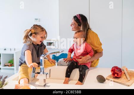 Madre e figlie allegre che giocano con acqua nel lavello della cucina a casa Foto Stock