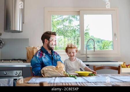 Felice padre e figlio godendo mentre si siedono al tavolo da pranzo con piselli verdi in cucina Foto Stock