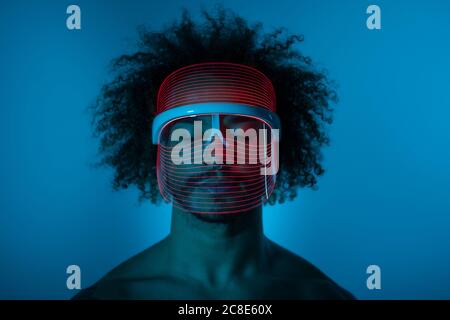 Giovane uomo con gli occhi chiusi indossando una maschera LED rossa contro parete a casa Foto Stock