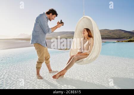 Uomo che prende foto di donna seduta in sedia pensile sopra piscina Foto Stock