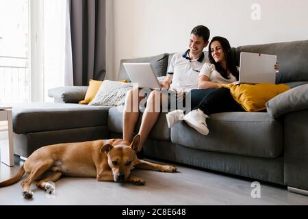 Accoppia usando i computer portatili sul divano vicino al cane a casa Foto Stock