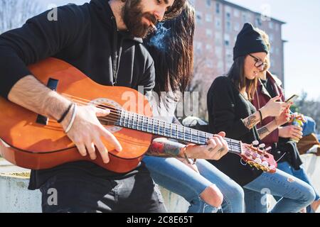 Uomo che fuma la sigaretta suonando la chitarra mentre si rilassa con gli amici città Foto Stock