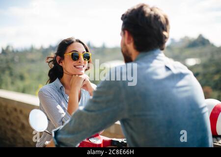 Donna sorridente con occhiali da sole che parla con il ragazzo seduto su Vespa Foto Stock