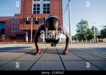 Atleta femminile con testina rasata che ascolta la musica attraverso le cuffie mentre fare push-up in strada in città Foto Stock