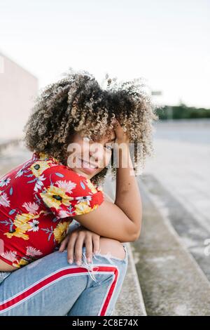 Felice giovane donna seduta con la mano in capelli sui gradini durante il giorno di sole Foto Stock