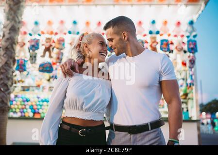 Ragazzo romantico guardando donna felice mentre si è in piedi contro stalla al parco divertimenti Foto Stock