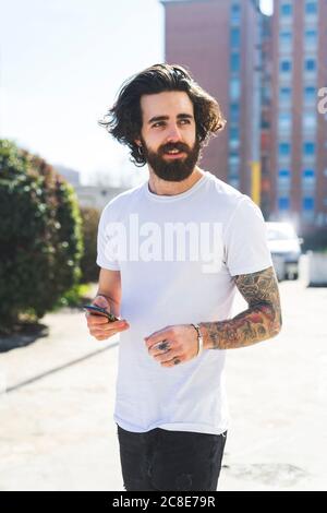 Giovane uomo bearded usando lo smartphone mentre camminando sulla strada durante il giorno di sole Foto Stock