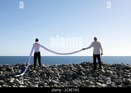 Donna con lunghe braccia artificiali che tengono la mano dell'uomo su ciottoli sulla spiaggia contro il cielo limpido Foto Stock