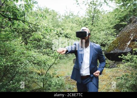 Uomo d'affari che pratica le arti marziali mentre osservano con simulatore di realtà virtuale contro gli alberi nella foresta Foto Stock