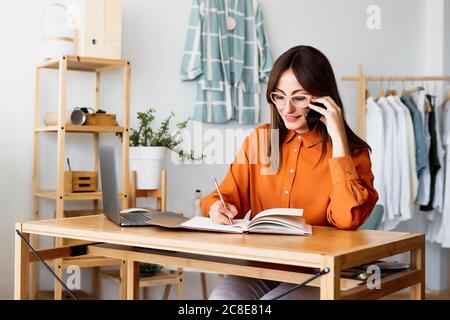 Donna stilista di moda che lavora a casa seduta alla scrivania prendendo note Foto Stock