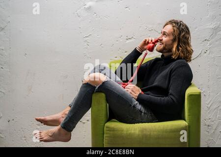 Uomo sorridente che parla al telefono mentre si siede sulla poltrona contro parete a casa Foto Stock