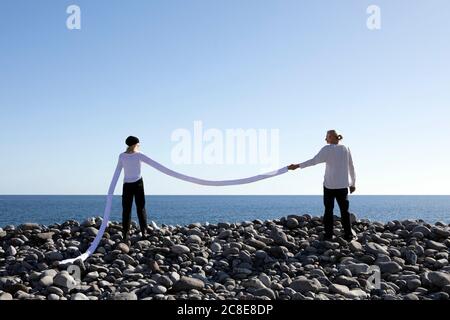 Donna con lunghe braccia artificiali che tengono la mano dell'uomo in spiaggia contro il cielo limpido Foto Stock