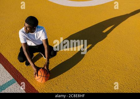 Premuroso giovane uomo che tiene il basket accovacciato sul campo sportivo durante giorno di sole Foto Stock