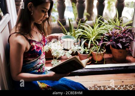 Donna che tiene la tazza mentre legge il libro dal balcone a casa Foto Stock