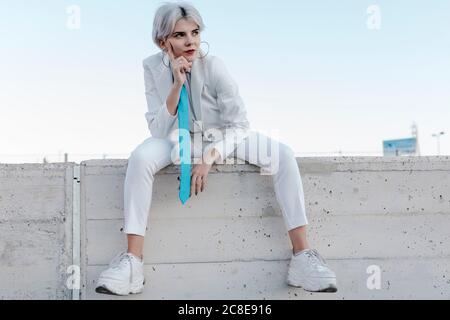 Donna giovane premurosa che indossa un abito elegante seduto su una parete di contenimento contro il cielo limpido Foto Stock