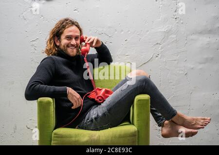 Uomo sorridente che parla al telefono mentre si rilassa sulla poltrona contro parete Foto Stock