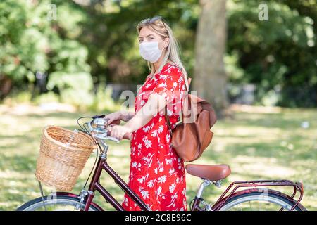 Donna mid adult che indossa la maschera mentre si è in piedi con la bicicletta dentro parco pubblico Foto Stock