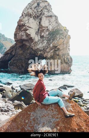 Giovane donna spensierata che scuotono la testa mentre si siede sulla roccia a Ursa Beach, Portogallo Foto Stock