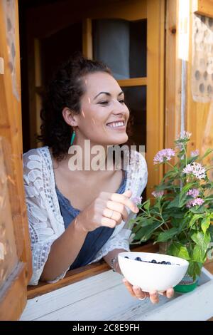 Donna felice che tiene i mirtilli in ciotola mentre sbircia attraverso la finestra di cottage Foto Stock