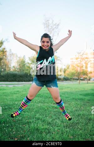 Adolescente ragazza con sindrome di Down indossando 80's colorful make-up e. i vestiti saltano sul prato Foto Stock