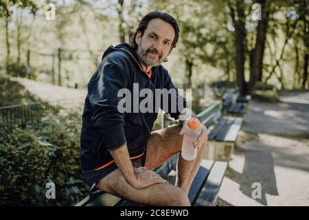 Uomo maturo sicuro che tiene la bottiglia d'acqua mentre si siede sul banco nel parco Foto Stock