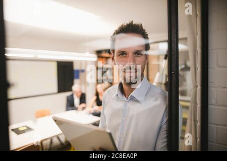 Uomo d'affari con tablet digitale, che si affaccia dalla finestra dell'ufficio Foto Stock